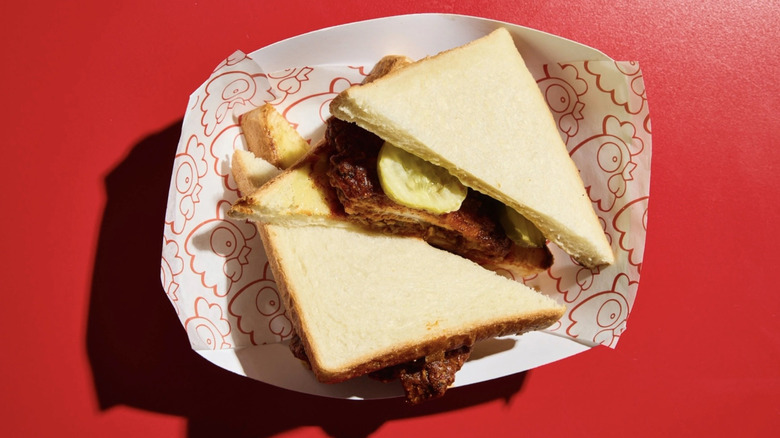 A fried chicken sandwich on white bread with pickles in a takeout container on a red background