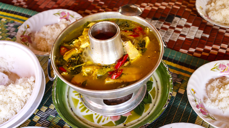 A pot of somlor machu soup on a table with plates of rice