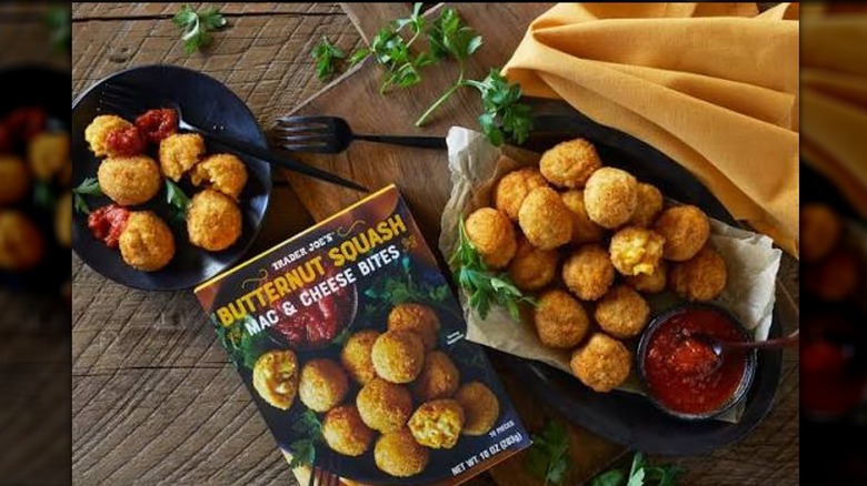 Box of Trader Joe's Butternut Squash Mac & Cheese Bites on a wooden table next to plates of cooked bites with sauce
