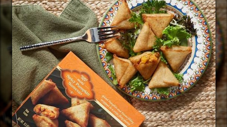 Plate of Trader Joe's Mini Spicy Pumpkin Samoas on top of greens next to the product's box on a woven placemat