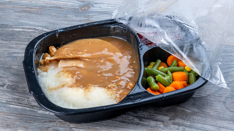 A turkey TV dinner with vegetables with lid halfway peeled off on wood surface.