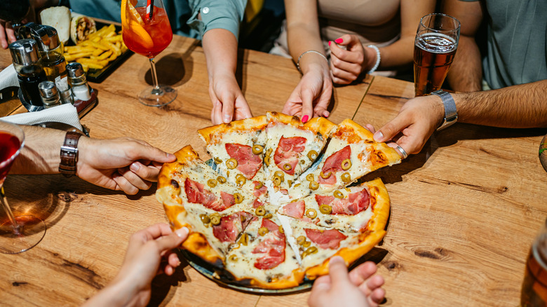 a group of people around a table taking a slice from a pizza pie