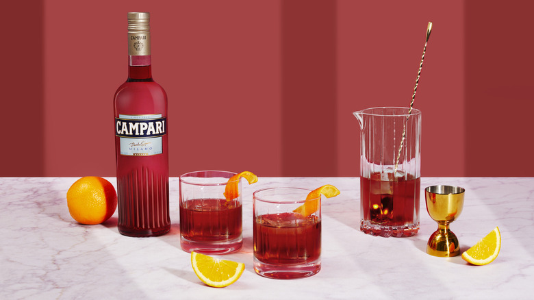 Negroni cocktails on a marble countertop next to a bottle of Campari, bartending tools, an orange, and orange slices
