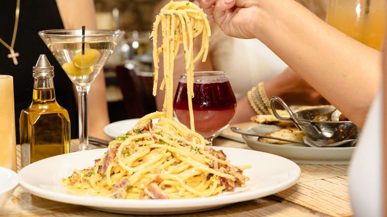 Person eating a plate of spaghetti alla carbonara and drinking a martini with olives
