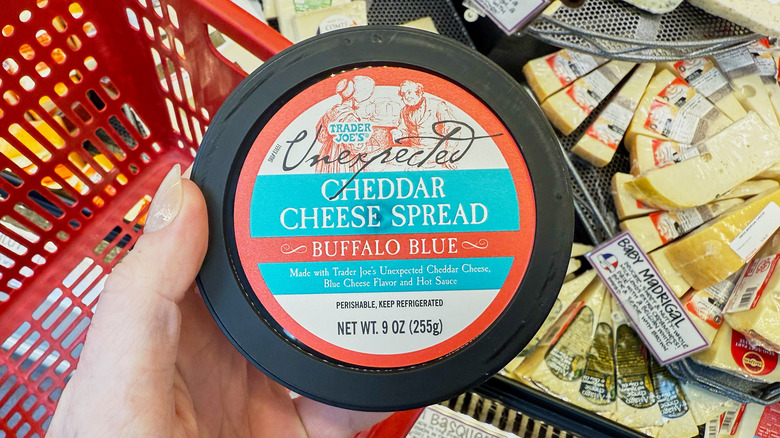 A hand holding a container of cheddar cheese spread in the cheese aisle at Trader Joe's