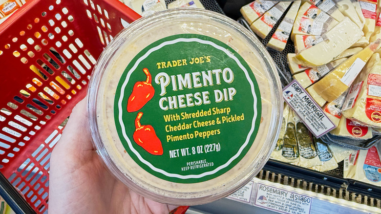 A hand holding a container of pimento cheese dip in the cheese aisle at Trader Joe's