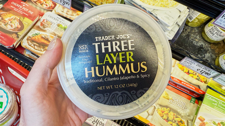 A hand holding a container of Trader Joe's Three Layer Hummus