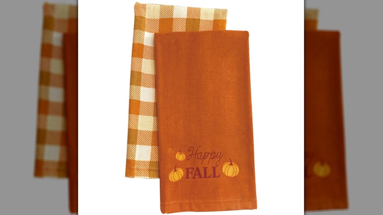 Two kitchen tea towels, one with a plaid orange design, the other with text reading "Happy Fall" with pumpkin decorations and an orange background.