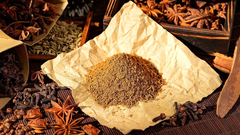 A mound of Chinese five-spice powder on a square piece of brown parchment paper surrounded by whole herbs like star anise, clove, and fennel