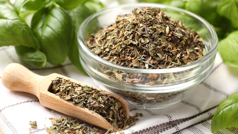 Dried basil in a small clear bowl and in a wooden rectangular scoop surrounded by fresh basil