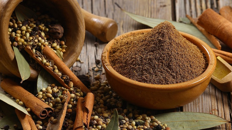 Garam masala powder in a tan rimmed bowl next a wooden container on its side with whole, spilled herbs