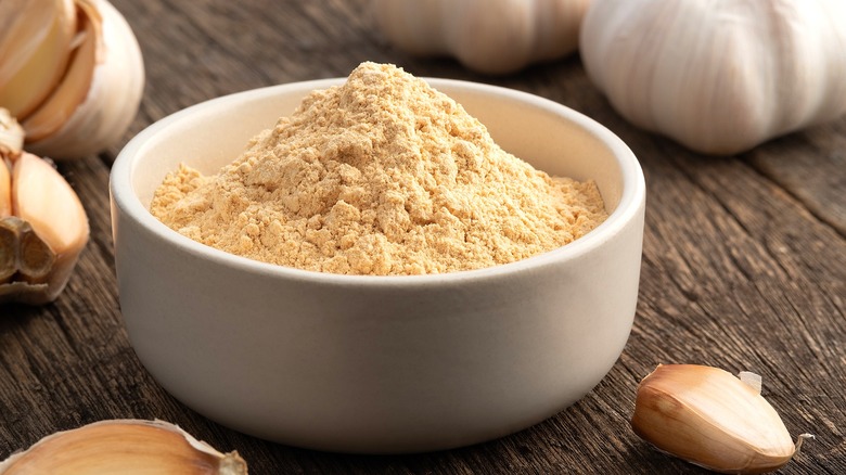 Garlic powder in a white bowl surrounded by whole cloves of unpeeled garlic