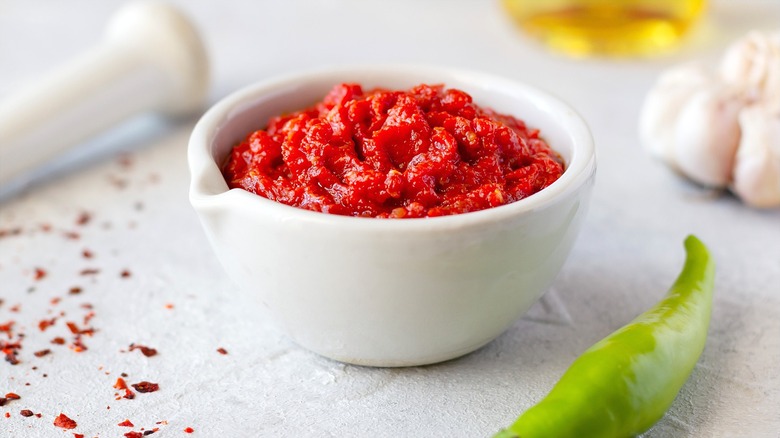 Harissa paste in a small white bowl surrounded by a head of garlic, one green pepper, and loose red pepper flakes