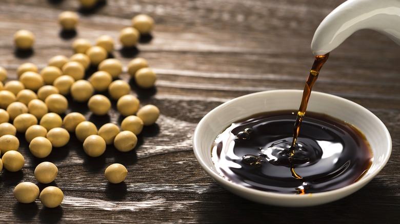 Soy sauce being poured into a small white shallow bowl next to whole soy beans