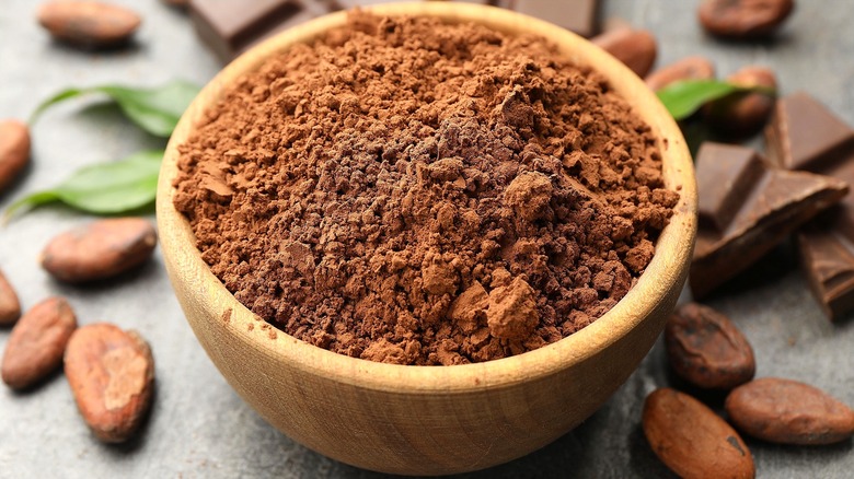Cocoa powder in a wooden bowl surrounded by cocoa beans, whole pieces of chocolate, and green leaves