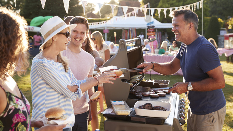 Man serving hot dogs and hamburgers from behind a grill to a line of people at a cookout.