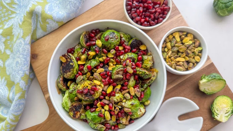 Bowl of Brussels sprouts with pomegranate seeds and pistachio on a wooden cutting board.