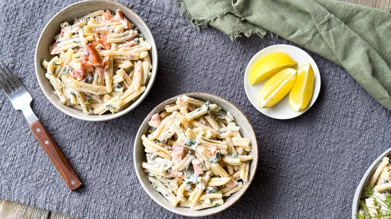 Pasta salad in bowls next to a plate of lemon wedges on a blue tablecloth.