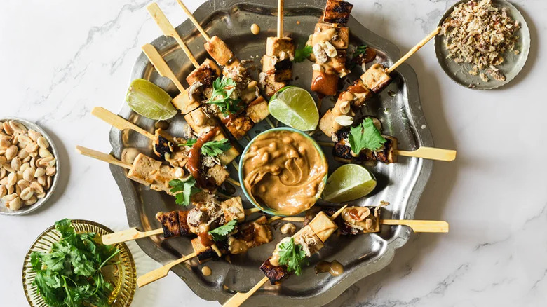 Tofu skewers with peanut sauce arranged on a silver plate.