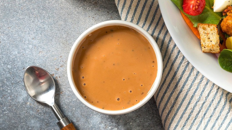 Balsamic salad dressing in a small white bowl next to a plate of salad and a metal spoon