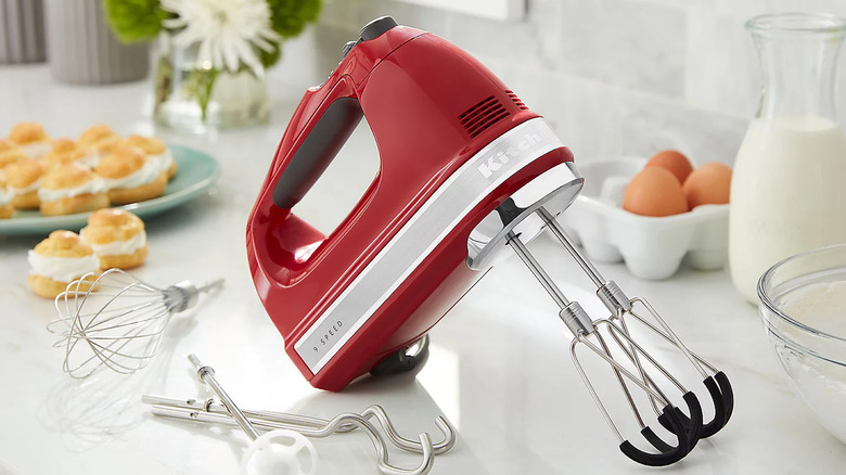 A red KitchenAid 9-speed digital hand mixer on a kitchen counter with cream puffs, eggs, and milk in the background