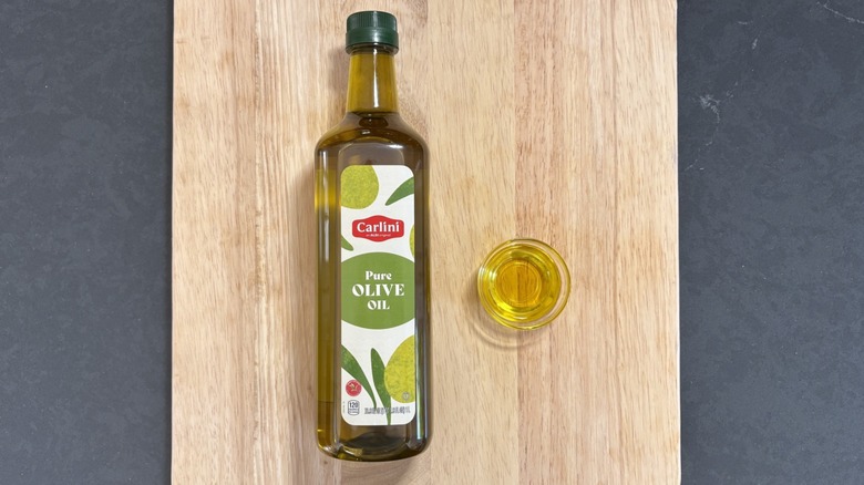 Bottle of Carlini Olive Oil with small glass dish on wooden cutting board and black counter