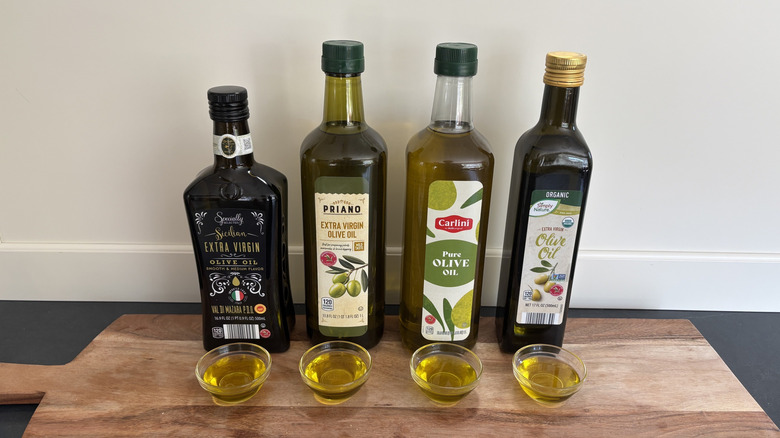 Four bottles of Aldi olive oil brands and small bowls of olive oil on cutting board