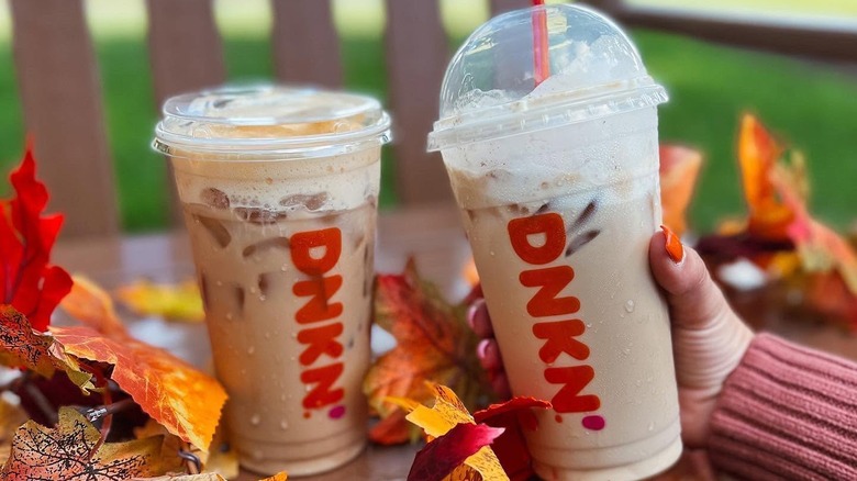 One Dunkin' iced latte sitting on a park bench, another being held next to it, both surrounded by colorful fall leaves.