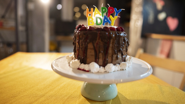 Chocolate birthday cake with "happy bday" candles on a white cake stand