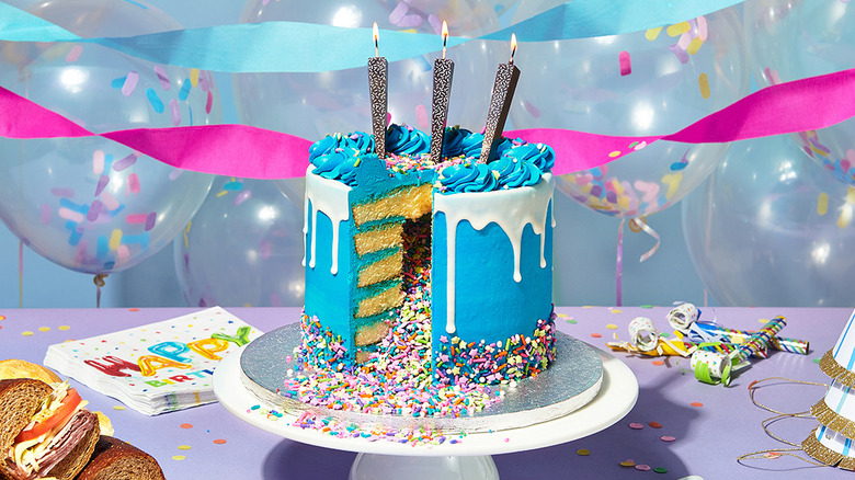 A multi-layered blue cake with white drizzle and sprinkles, with balloons and streamers in the background.