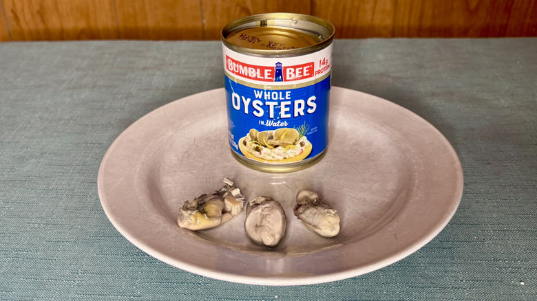 A can of Bumble Bee Whole Oysters in Water behind three individual oysters, on a plate