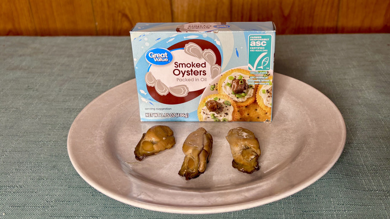 A box of Great Value Smoked Oysters on a plate, sitting behind three individual oysters