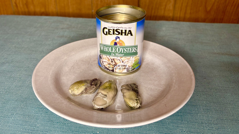 A can of Geisha Whole Oysters in Water rests on a plate behind three individual oysters
