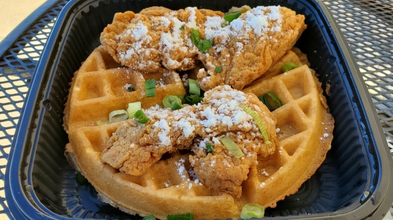 Chicken and waffles with powdered sugar and chives from Scramblers in a black takeout container.