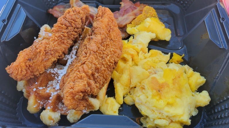 Chicken strips and a waffle with scrambled eggs and bacon from Denny's in a black takeout container.