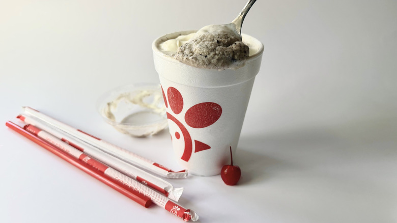 Chick-fil-A's Cookies & Cream Milkshake with a spoon inserted into the cup next to a cherry, straws, and the lid