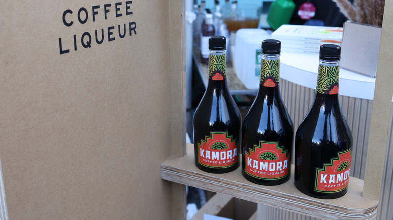 Three bottles of Kamora Coffee Liqueur on a display case, the words Coffee Liqueur printed in a sans serif font on the wall beside them