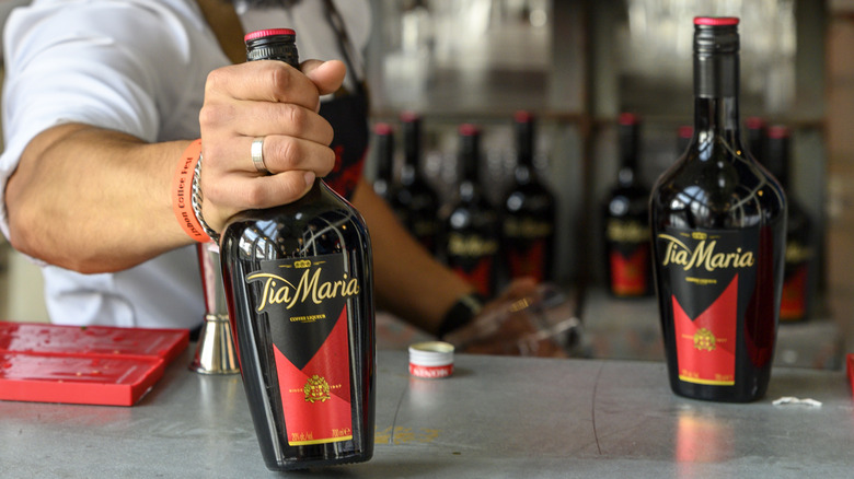 A muscular bartender holds a bottle of Tia Maria coffee liqueur across a slate gray bar, another bottle of Tia Maria is in the near background, several bottles are seen in the unfocused background