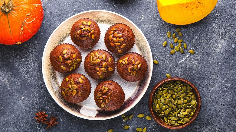 Pumpkin muffins with pumpkin seeds on a round platter surrounded by cut squash and a small whole pumpkin