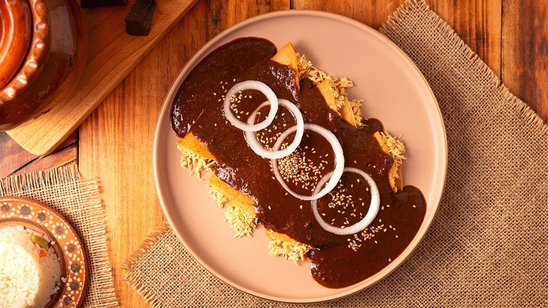 Top-down view of enchiladas with shredded chicken covered in mole sauce, raw white onion slices, and sesame seeds