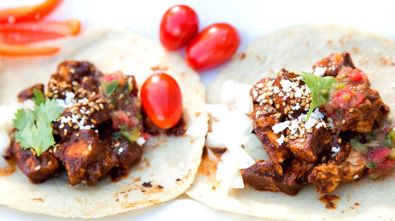 Two open-faced flour tortillas filled with cubed chicken in mole, cilantro, cherry tomatoes, and sesame seeds