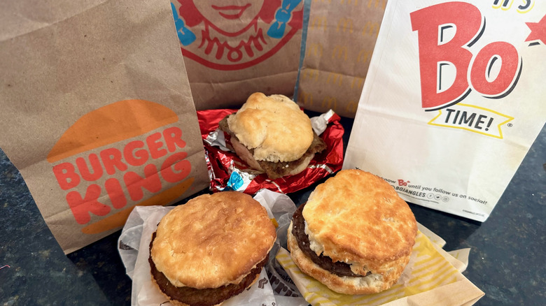 three fast food sausage biscuits on their wrappers with takeout bags behind them