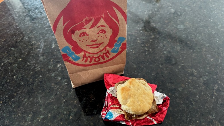 Wendy's sausage biscuit on its wrapper with takeout bag behind it