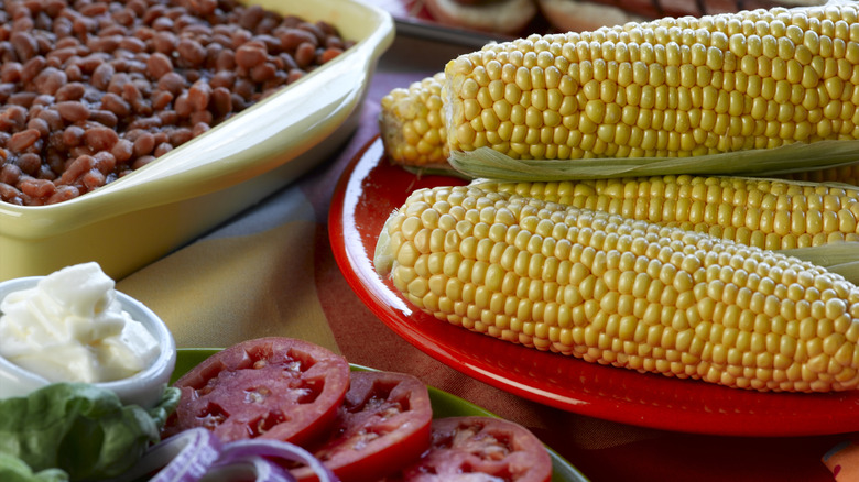 Barbecue sides like corn on the cob, beans, and veggies on plates.