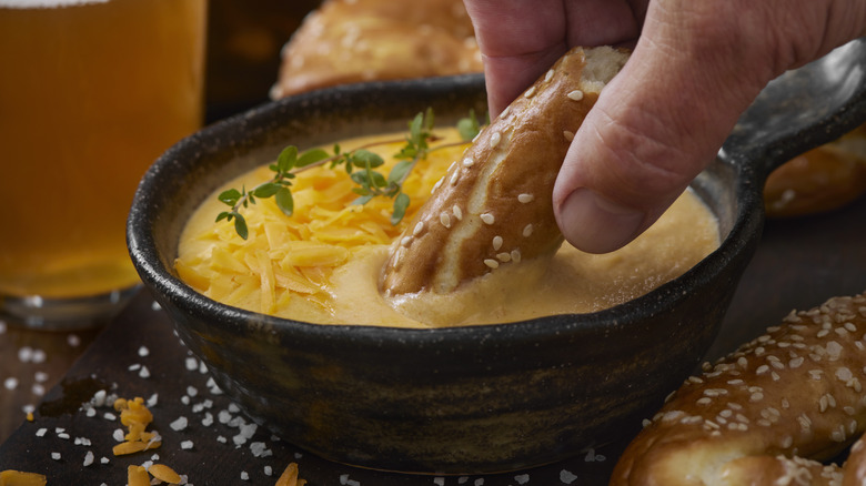 A hand dipping a sesame-seed covered soft pretzel piece into a small skillet of beer cheese
