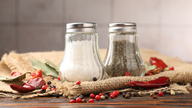 Salt and pepper shakers on burlap and a wood surface surrounded by peppercorns and dried chiles