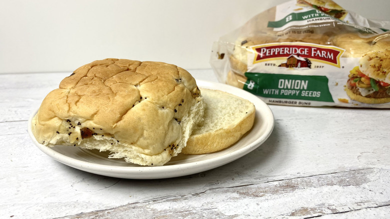 A Pepperidge Farm Onion with Poppy Seeds Hamburger Bun on a white plate in front of its packaging on a white surface.