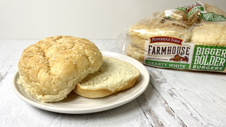 A Pepperidge Farm Farmhouse Hearty White Hamburger Bun on a white plate in front of its packaging on a white surface.