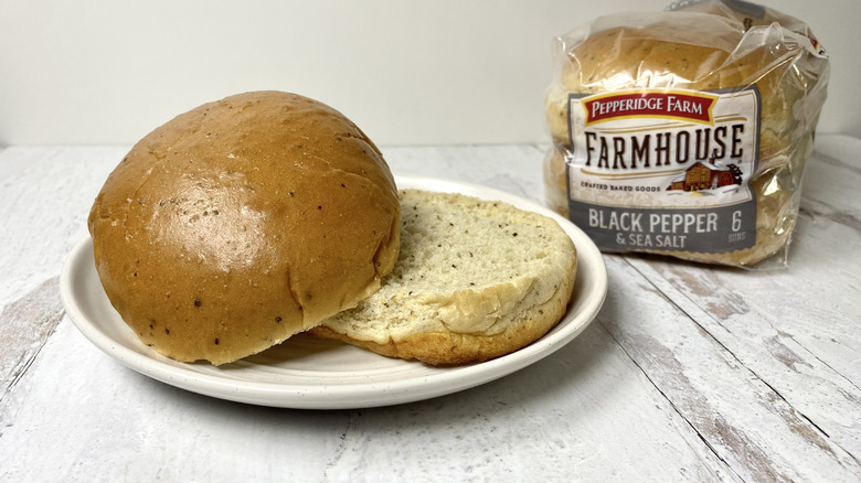 A Pepperidge Farm Farmhouse Black Pepper & Sea Salt Hamburger Bun on a white plate in front of its packaging on a white surface.