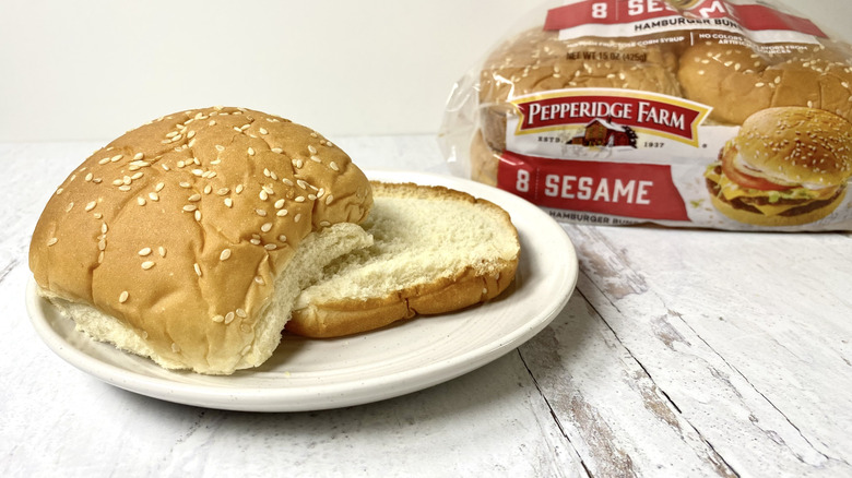 A Pepperidge Farm Sesame Hamburger Bun on a white plate in front of its packaging on a white surface.